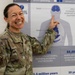 A female military service member in U.S. Army OCP (Operational Camouflage Pattern) uniform is smiling broadly while pointing at an informational poster about the effects of alcohol on the human body. Her name tape reads 'GLOVELTER' and she wears a unit patch identified as '376th OP Medical Readiness SQ' with a caduceus symbol. The poster behind her displays statistics and anatomical diagrams related to alcohol-induced health risks and deaths.