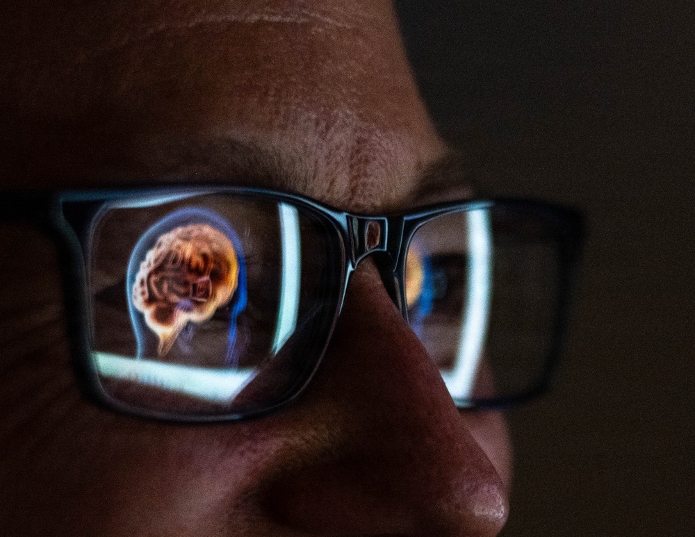 A close-up photograph of a person wearing dark-framed rectangular glasses in a dimly lit environment. Reflected in the left lens is a glowing digital or medical illustration of a human brain inside a head silhouette, suggesting the person is viewing a screen displaying neurological or medical imaging content. The right lens shows bright light streaks, likely from the same screen source.