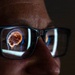 A close-up photograph of a person wearing dark-framed rectangular glasses in a dimly lit environment. Reflected in the left lens is a glowing digital or medical illustration of a human brain inside a head silhouette, suggesting the person is viewing a screen displaying neurological or medical imaging content. The right lens shows bright light streaks, likely from the same screen source.