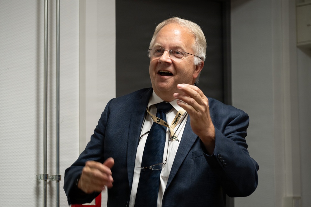 A middle-aged to older man wearing a navy blue suit, white dress shirt, and navy tie is captured mid-speech or presentation. He is gesturing with both hands expressively and wearing glasses on a lanyard around his neck. A gold lanyard with text reading 'GO ARMY' is visible around his neck.