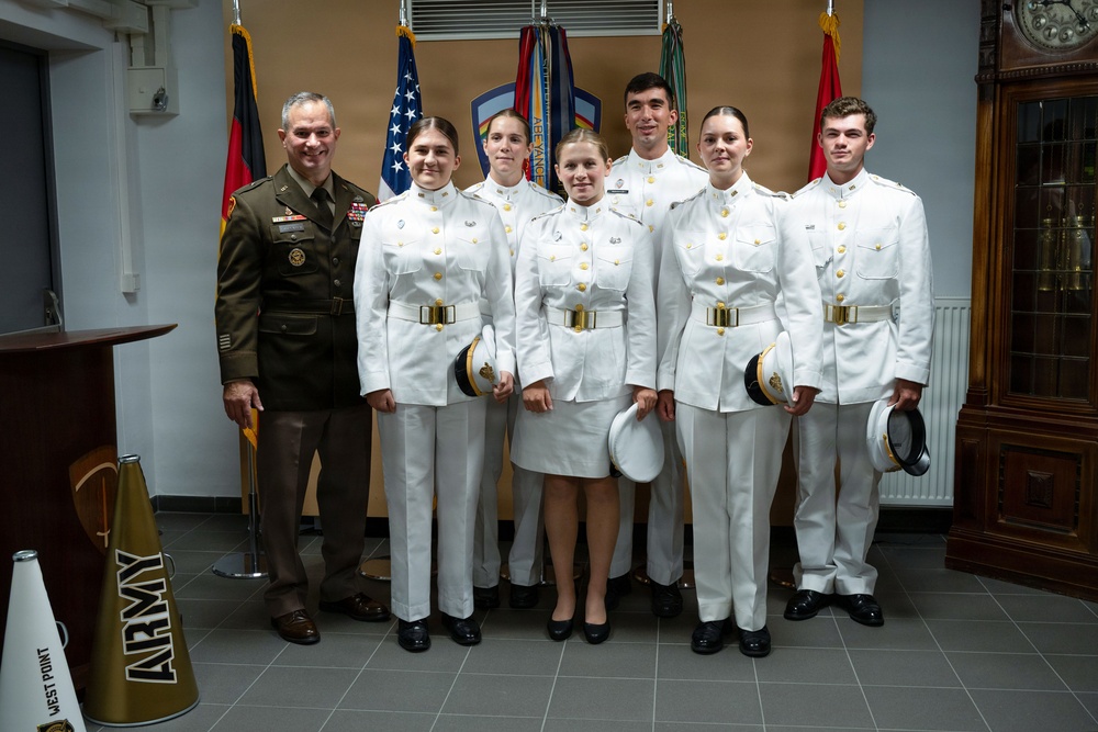 A group of seven individuals pose for a formal photograph in what appears to be an official military setting. One person on the left wears a U.S. Army dress uniform (olive/green), while five others wear white military dress uniforms consistent with West Point cadet formal attire. A megaphone with 'ARMY WEST POINT' branding is visible in the lower left corner.