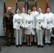 A group of seven individuals pose for a formal photograph in what appears to be an official military setting. One person on the left wears a U.S. Army dress uniform (olive/green), while five others wear white military dress uniforms consistent with West Point cadet formal attire. A megaphone with 'ARMY WEST POINT' branding is visible in the lower left corner.