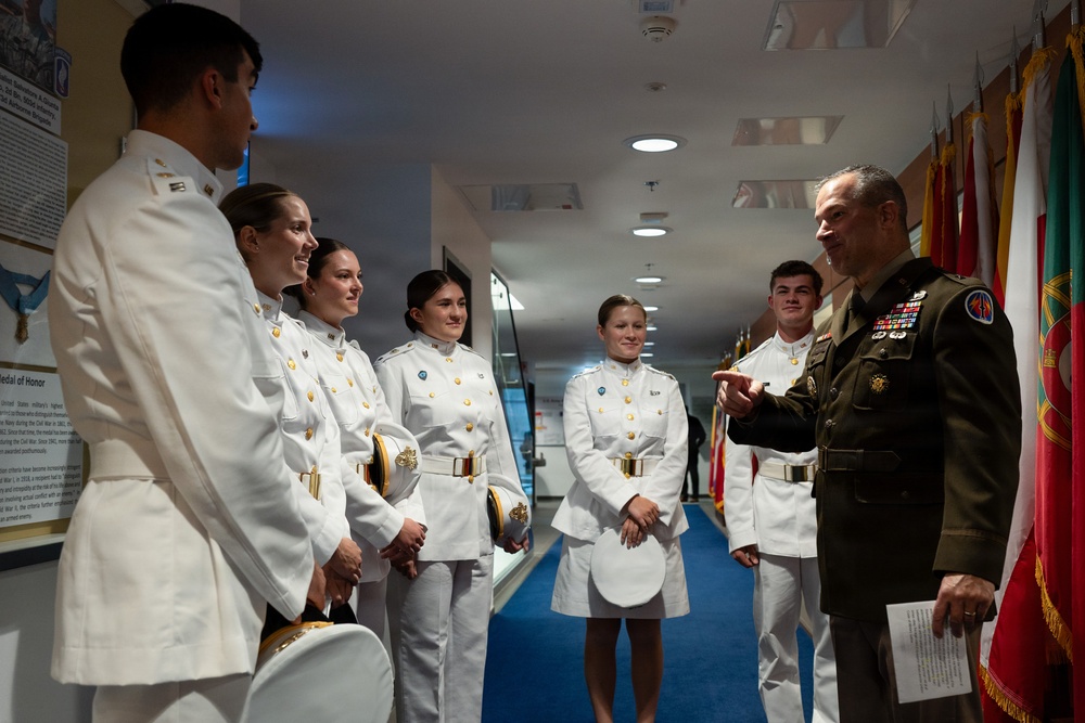 A senior military officer in olive green dress uniform with extensive medal ribbons is speaking with a group of cadets or midshipmen dressed in white dress uniforms. The setting appears to be an indoor military facility or museum corridor, with national and military flags displayed along the right wall. The interaction appears to be an informal briefing or meet-and-greet.