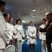 A senior military officer in olive green dress uniform with extensive medal ribbons is speaking with a group of cadets or midshipmen dressed in white dress uniforms. The setting appears to be an indoor military facility or museum corridor, with national and military flags displayed along the right wall. The interaction appears to be an informal briefing or meet-and-greet.