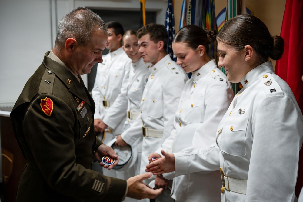 A U.S. military officer in olive green dress uniform is presenting items, appearing to be coins or patches, to cadets or junior officers dressed in white dress uniforms. The ceremony appears to be a formal recognition or commissioning event taking place indoors with military flags visible in the background. Multiple recipients are lined up awaiting their turn in what appears to be an official military ceremony.