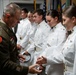 A U.S. military officer in olive green dress uniform is presenting items, appearing to be coins or patches, to cadets or junior officers dressed in white dress uniforms. The ceremony appears to be a formal recognition or commissioning event taking place indoors with military flags visible in the background. Multiple recipients are lined up awaiting their turn in what appears to be an official military ceremony.