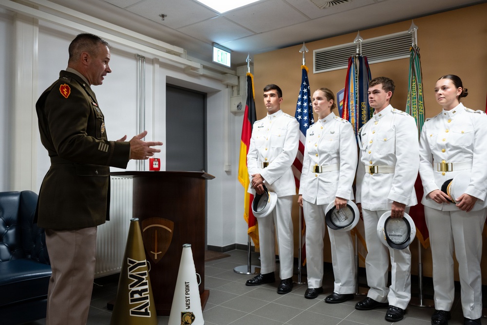 A military officer in dress uniform addresses four cadets in white dress uniforms at what appears to be a formal ceremony. The setting includes multiple flags including the American and German flags, and items branded with 'Army' and 'West Point' are visible near the podium. This appears to be a West Point Military Academy event held at a location in Germany.