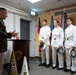A military officer in dress uniform addresses four cadets in white dress uniforms at what appears to be a formal ceremony. The setting includes multiple flags including the American and German flags, and items branded with 'Army' and 'West Point' are visible near the podium. This appears to be a West Point Military Academy event held at a location in Germany.