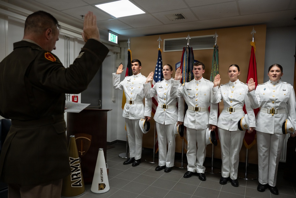 A military commissioning or oath-of-office ceremony is taking place indoors. Five cadets in white dress uniforms are raising their right hands while a senior officer in olive green uniform administers the oath from a podium. The setting includes multiple military flags and West Point memorabilia, indicating this is a U.S. Military Academy (West Point) event.
