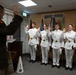 A military commissioning or oath-of-office ceremony is taking place indoors. Five cadets in white dress uniforms are raising their right hands while a senior officer in olive green uniform administers the oath from a podium. The setting includes multiple military flags and West Point memorabilia, indicating this is a U.S. Military Academy (West Point) event.