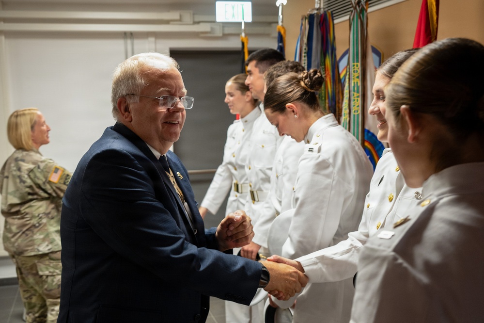 A civilian man in a dark blue suit is shaking hands with military personnel dressed in white dress uniforms during what appears to be a formal ceremony or reception. Several cadets or officers are lined up in a receiving line formation. In the background, a person in Army camouflage (OCP) uniform and multiple military unit flags/colors are visible.