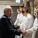 A civilian man in a dark blue suit is shaking hands with military personnel dressed in white dress uniforms during what appears to be a formal ceremony or reception. Several cadets or officers are lined up in a receiving line formation. In the background, a person in Army camouflage (OCP) uniform and multiple military unit flags/colors are visible.