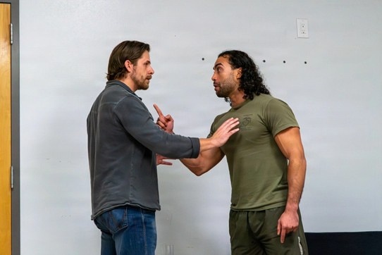 Two men are engaged in what appears to be a tense confrontation or instructional demonstration in an indoor setting. The man on the left is pointing a finger while the man on the right has his hand extended in a stopping or deflecting gesture. The interaction suggests either a conflict scenario, self-defense training, or a staged dramatic scene.