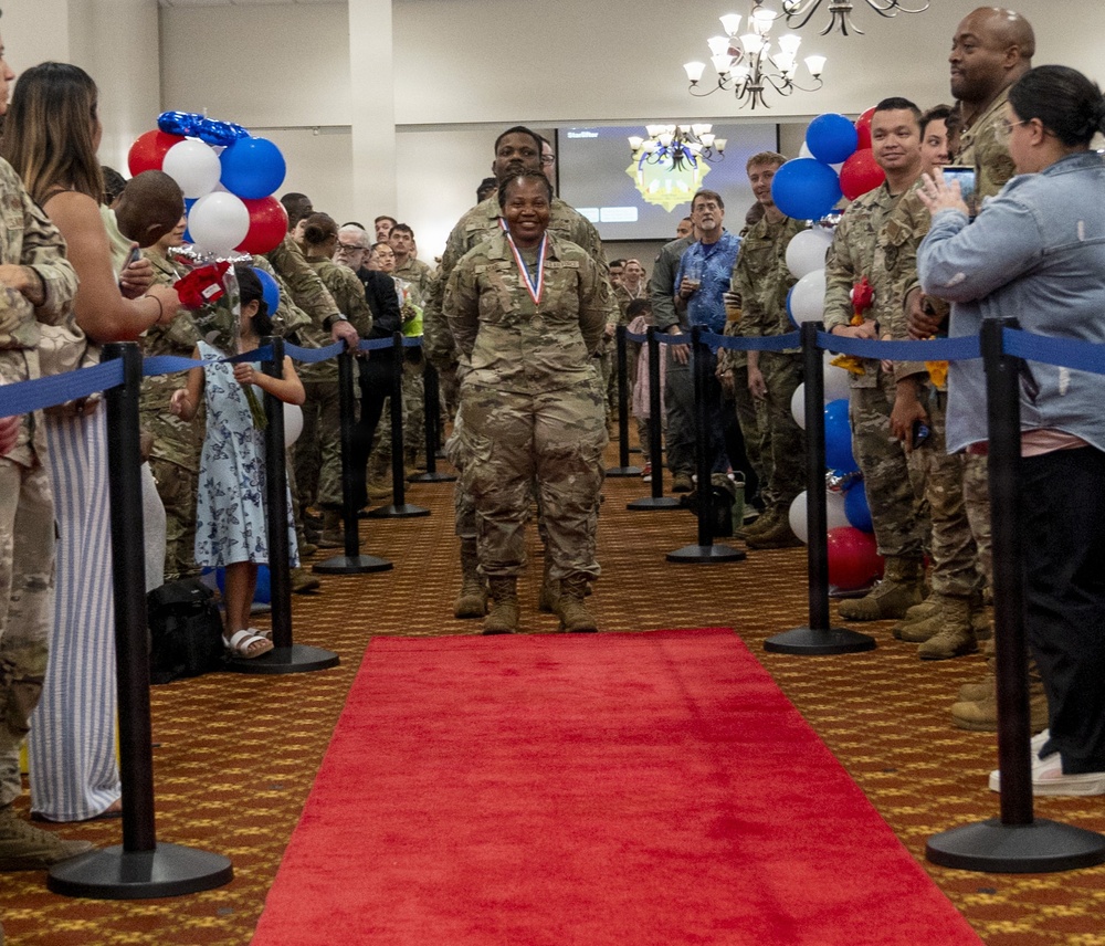 A military ceremony taking place in an indoor venue where a female soldier wearing a medal walks down a red carpet flanked by stanchions with blue ropes. She is surrounded by fellow soldiers in camouflage uniforms and civilians who appear to be applauding and celebrating. The event is decorated with red, white, and blue balloons, suggesting a patriotic military recognition or welcome-home ceremony.