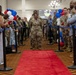 A military ceremony taking place in an indoor venue where a female soldier wearing a medal walks down a red carpet flanked by stanchions with blue ropes. She is surrounded by fellow soldiers in camouflage uniforms and civilians who appear to be applauding and celebrating. The event is decorated with red, white, and blue balloons, suggesting a patriotic military recognition or welcome-home ceremony.