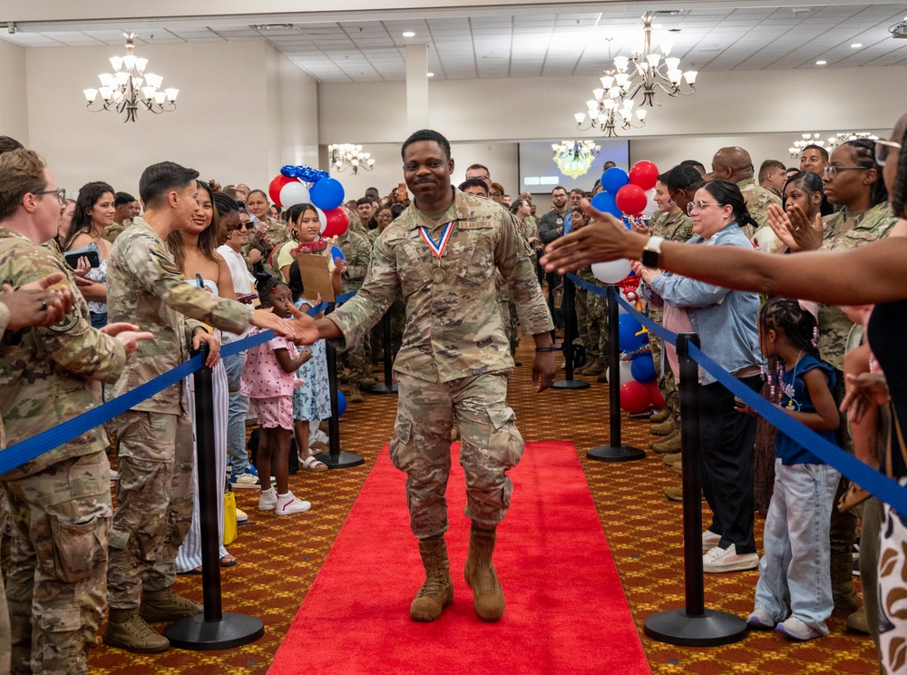 A military service member wearing a medal walks down a red carpet through a crowd of fellow service members, families, and civilians in what appears to be an indoor ceremony or recognition event. The atmosphere is celebratory, with people on both sides reaching out to high-five or shake hands with the honoree. Red, white, and blue balloons decorate the venue, suggesting a patriotic or military celebration.
