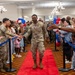 A military service member wearing a medal walks down a red carpet through a crowd of fellow service members, families, and civilians in what appears to be an indoor ceremony or recognition event. The atmosphere is celebratory, with people on both sides reaching out to high-five or shake hands with the honoree. Red, white, and blue balloons decorate the venue, suggesting a patriotic or military celebration.
