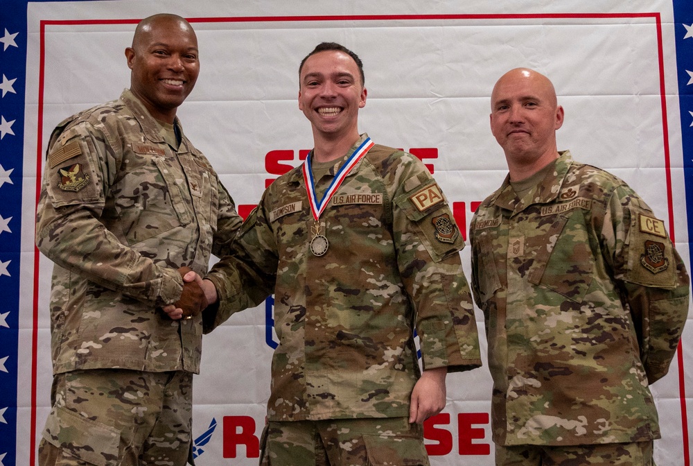 Three U.S. Air Force personnel pose for a photo at what appears to be an award ceremony. The center individual, identified by name tape as 'Thompson,' is wearing a red, white, and blue medal/medallion on a ribbon around their neck. The person on the left is shaking hands with Thompson while all three smile for the camera.