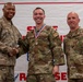 Three U.S. Air Force personnel pose for a photo at what appears to be an award ceremony. The center individual, identified by name tape as 'Thompson,' is wearing a red, white, and blue medal/medallion on a ribbon around their neck. The person on the left is shaking hands with Thompson while all three smile for the camera.