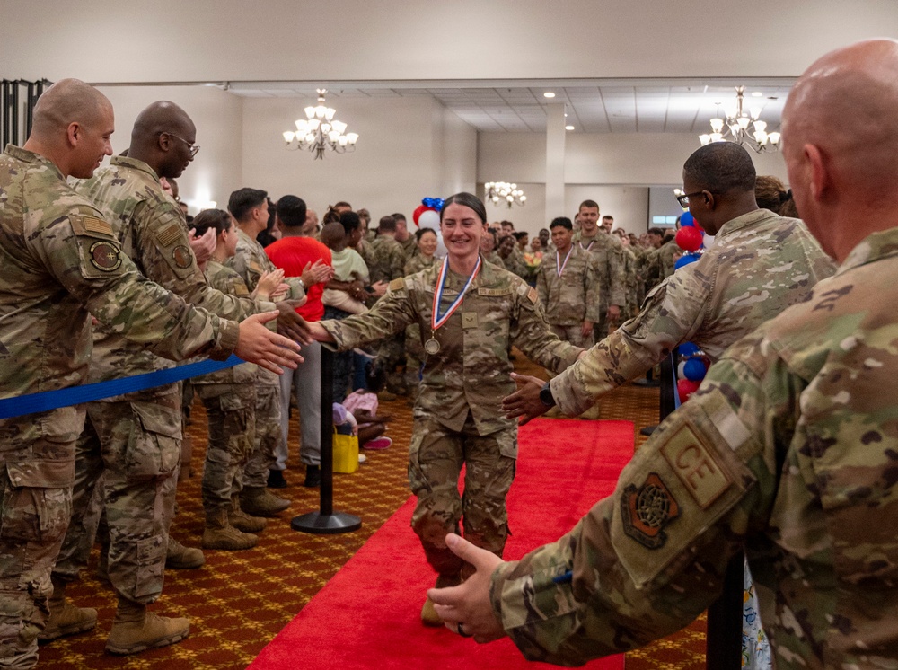 A military service member wearing a medal walks down a red carpet through a gauntlet of fellow soldiers applauding and offering handshakes in what appears to be an indoor ceremony hall. The central figure, a female airman in OCP (Operational Camouflage Pattern) uniform, is being celebrated and congratulated by peers lined up on both sides. Red, white, and blue balloons are visible in the background, suggesting a patriotic or awards ceremony event.