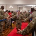 A military service member wearing a medal walks down a red carpet through a gauntlet of fellow soldiers applauding and offering handshakes in what appears to be an indoor ceremony hall. The central figure, a female airman in OCP (Operational Camouflage Pattern) uniform, is being celebrated and congratulated by peers lined up on both sides. Red, white, and blue balloons are visible in the background, suggesting a patriotic or awards ceremony event.