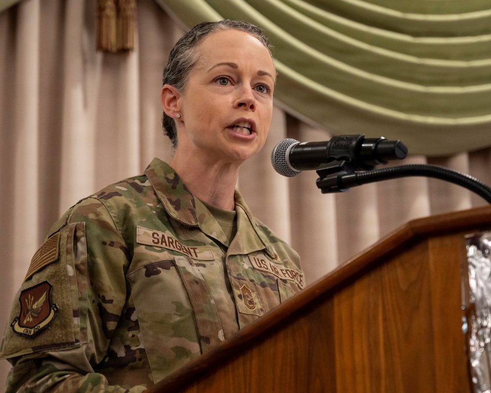 A U.S. Air Force service member named Sargent, wearing OCP (Operational Camouflage Pattern) uniform, is speaking at a podium with a microphone. The name tape reads 'SARGENT' and the branch tape reads 'US AIR FORCE,' with a Master Sergeant rank insignia visible on the chest. The setting appears to be a formal indoor military event or ceremony.