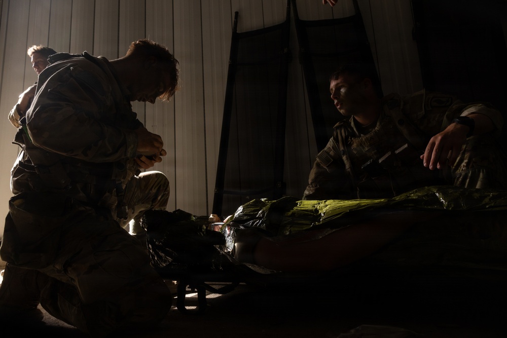 Military personnel in camouflage uniforms are gathered around what appears to be a litter or stretcher in a dimly lit indoor space. The scene appears to depict a tactical combat casualty care (TCCC) training exercise or medical treatment scenario. At least three individuals are visible, with one kneeling and actively working on the subject on the stretcher.