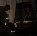 Military personnel in camouflage uniforms are gathered around what appears to be a litter or stretcher in a dimly lit indoor space. The scene appears to depict a tactical combat casualty care (TCCC) training exercise or medical treatment scenario. At least three individuals are visible, with one kneeling and actively working on the subject on the stretcher.