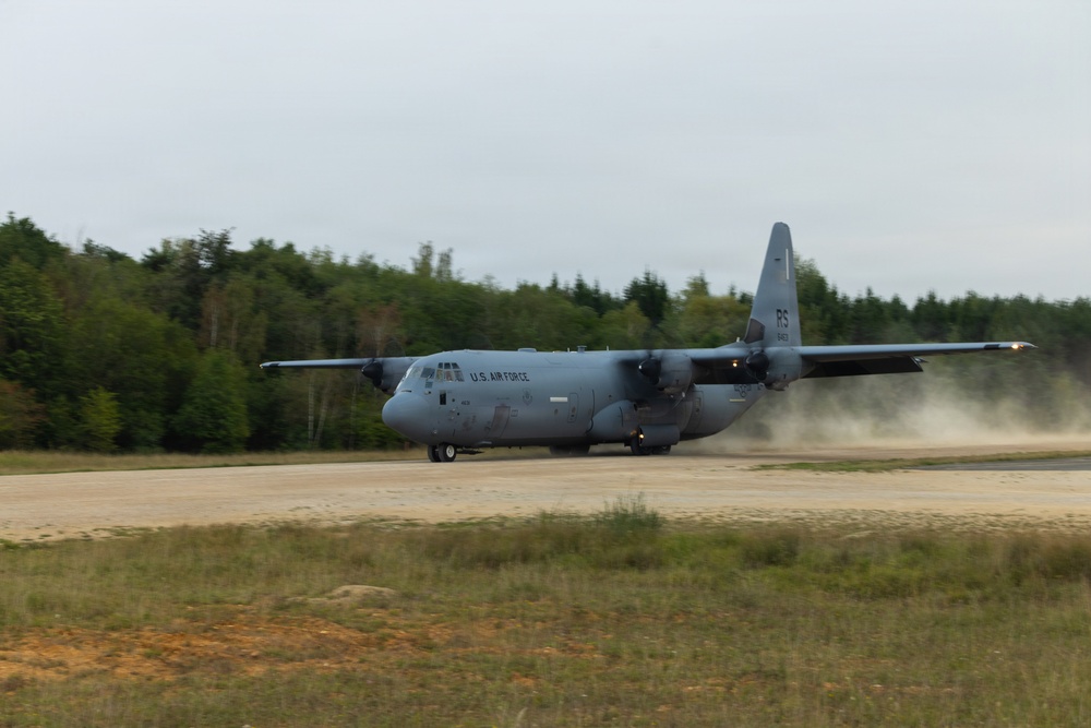 A U.S. Air Force C-130 Hercules military transport aircraft is taxiing or landing on a runway, kicking up a significant dust cloud behind it. The aircraft bears 'U.S. AIR FORCE' markings and tail code 'RS', indicating it is assigned to a unit based in Europe (Ramstein Air Base). The setting appears to be a semi-improved or austere airstrip surrounded by dense forest under overcast skies.