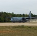 A U.S. Air Force C-130 Hercules military transport aircraft is taxiing or landing on a runway, kicking up a significant dust cloud behind it. The aircraft bears 'U.S. AIR FORCE' markings and tail code 'RS', indicating it is assigned to a unit based in Europe (Ramstein Air Base). The setting appears to be a semi-improved or austere airstrip surrounded by dense forest under overcast skies.
