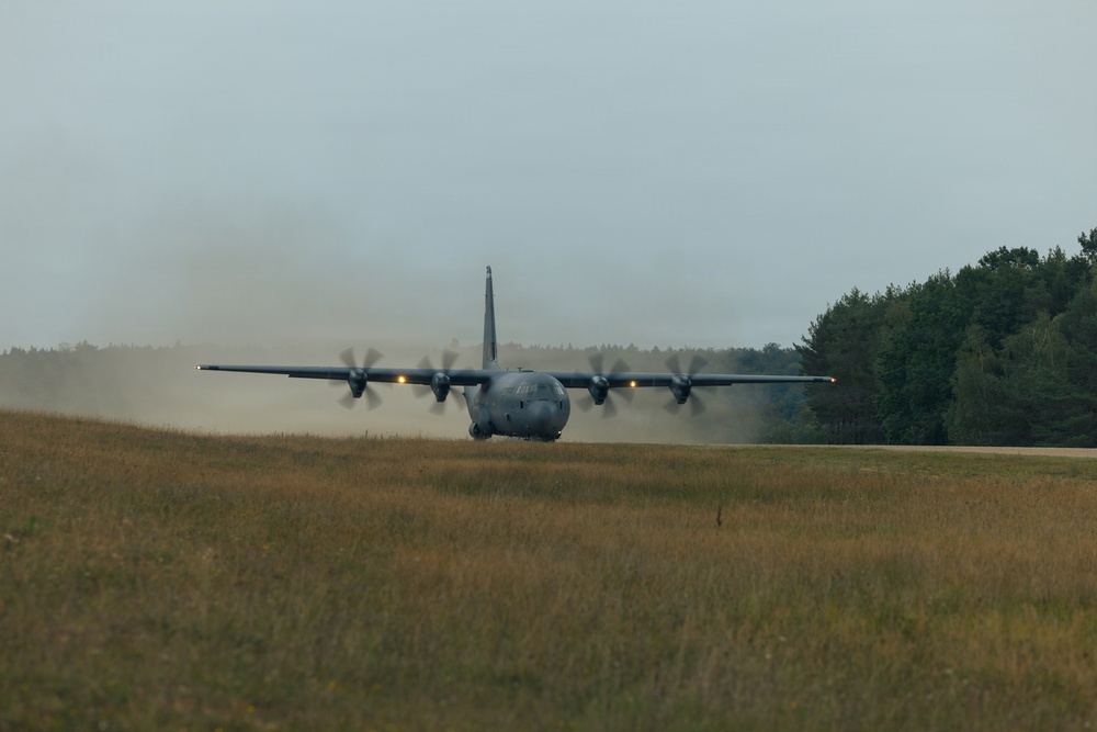 A large military turboprop transport aircraft, consistent with a C-130 Hercules variant, is operating on an unpaved or semi-prepared airstrip. The aircraft is generating a significant dust cloud behind it, suggesting it is either landing or performing a short-field takeoff roll. The setting appears to be a rural or semi-wooded area under overcast skies.