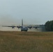 A large military turboprop transport aircraft, consistent with a C-130 Hercules variant, is operating on an unpaved or semi-prepared airstrip. The aircraft is generating a significant dust cloud behind it, suggesting it is either landing or performing a short-field takeoff roll. The setting appears to be a rural or semi-wooded area under overcast skies.