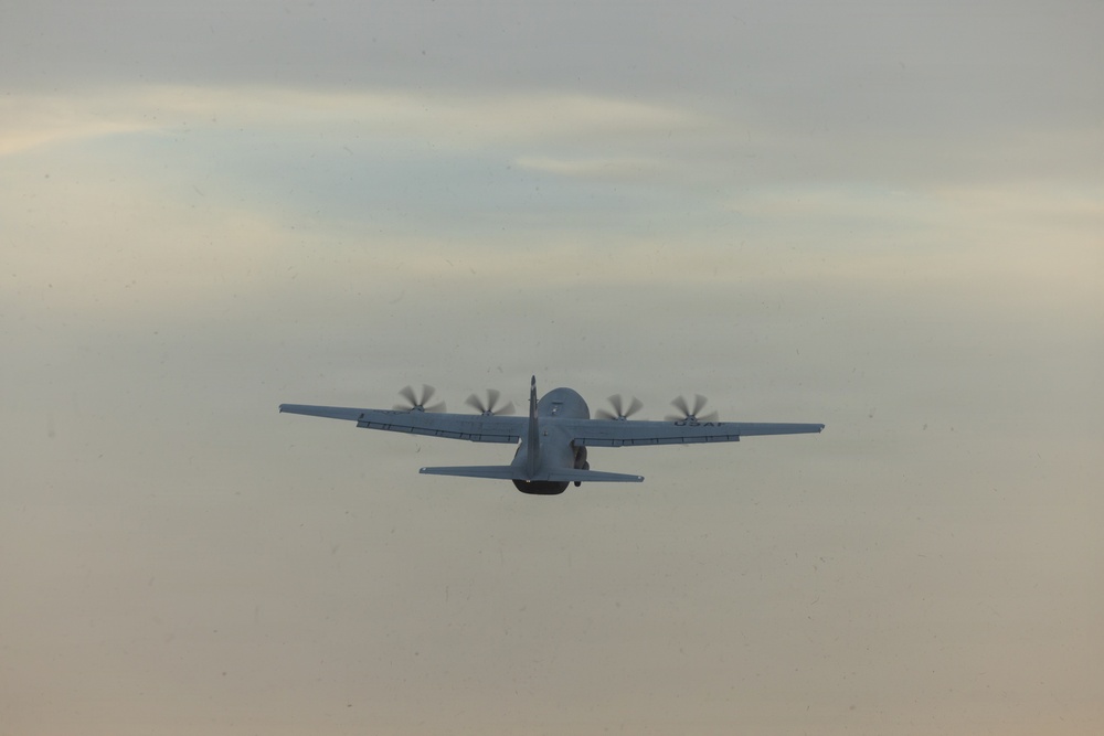 A four-engine turboprop military transport aircraft is photographed in flight from a rear perspective against an overcast, hazy sky. The aircraft appears to be a C-130 Hercules or similar variant, identifiable by its high-wing configuration and four propeller engines. All four propellers are visibly spinning, indicating active flight.