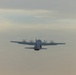 A four-engine turboprop military transport aircraft is photographed in flight from a rear perspective against an overcast, hazy sky. The aircraft appears to be a C-130 Hercules or similar variant, identifiable by its high-wing configuration and four propeller engines. All four propellers are visibly spinning, indicating active flight.