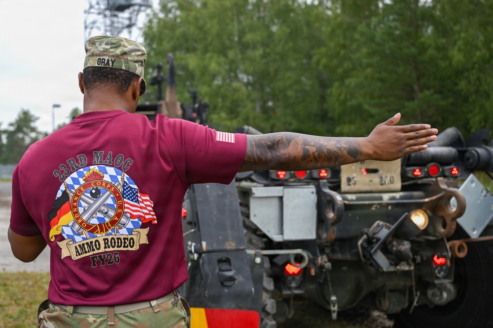 A U.S. Army soldier named Gray, wearing a maroon '23rd MAOC Ammo Rodeo FY26' t-shirt and multicam patrol cap, is directing a military vehicle with an outstretched arm. The soldier has visible tattoos on their right forearm and is wearing a small U.S. flag patch on the sleeve. The setting appears to be a military installation or training area with trees and overcast skies in the background.