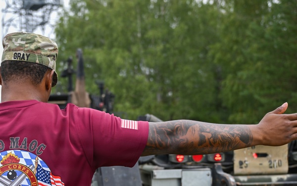 A U.S. Army soldier named Gray, wearing a maroon '23rd MAOC Ammo Rodeo FY26' t-shirt and multicam patrol cap, is directing a military vehicle with an outstretched arm. The soldier has visible tattoos on their right forearm and is wearing a small U.S. flag patch on the sleeve. The setting appears to be a military installation or training area with trees and overcast skies in the background.