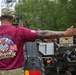 A U.S. Army soldier named Gray, wearing a maroon '23rd MAOC Ammo Rodeo FY26' t-shirt and multicam patrol cap, is directing a military vehicle with an outstretched arm. The soldier has visible tattoos on their right forearm and is wearing a small U.S. flag patch on the sleeve. The setting appears to be a military installation or training area with trees and overcast skies in the background.