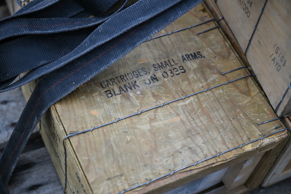 The image shows a weathered wooden ammunition crate stenciled with 'CARTRIDGES, SMALL ARMS, BLANK UN 0338', indicating it contains blank cartridges classified under UN hazardous materials code 0338. The crate is secured with wire banding and has dark nylon strapping draped over it. A second wooden crate is partially visible to the right with partial text markings.