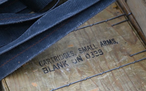 The image shows a weathered wooden ammunition crate stenciled with 'CARTRIDGES, SMALL ARMS, BLANK UN 0338', indicating it contains blank cartridges classified under UN hazardous materials code 0338. The crate is secured with wire banding and has dark nylon strapping draped over it. A second wooden crate is partially visible to the right with partial text markings.