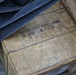 The image shows a weathered wooden ammunition crate stenciled with 'CARTRIDGES, SMALL ARMS, BLANK UN 0338', indicating it contains blank cartridges classified under UN hazardous materials code 0338. The crate is secured with wire banding and has dark nylon strapping draped over it. A second wooden crate is partially visible to the right with partial text markings.