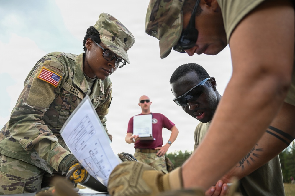 A group of U.S. Army soldiers in MultiCam (OCP) uniforms are gathered around a surface reviewing documents or maps during what appears to be a field training exercise. The soldier on the left, identified by the name tape 'LILLY,' is holding printed materials and appears to be coordinating with fellow soldiers. A third soldier in the background wearing a maroon t-shirt observes while holding a clipboard.