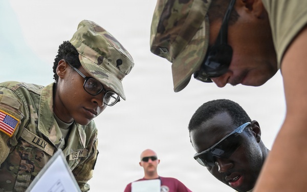 A group of U.S. Army soldiers in MultiCam (OCP) uniforms are gathered around a surface reviewing documents or maps during what appears to be a field training exercise. The soldier on the left, identified by the name tape 'LILLY,' is holding printed materials and appears to be coordinating with fellow soldiers. A third soldier in the background wearing a maroon t-shirt observes while holding a clipboard.