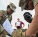 A group of U.S. Army soldiers in MultiCam (OCP) uniforms are gathered around a surface reviewing documents or maps during what appears to be a field training exercise. The soldier on the left, identified by the name tape 'LILLY,' is holding printed materials and appears to be coordinating with fellow soldiers. A third soldier in the background wearing a maroon t-shirt observes while holding a clipboard.