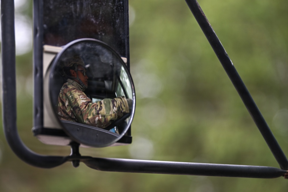 A person wearing military camouflage uniform and a cap is visible reflected in a vehicle's side mirror assembly. The reflection shows the individual seated inside a vehicle, appearing to be driving or operating it. The image is composed as a close-up of the mirror hardware against a blurred green background.