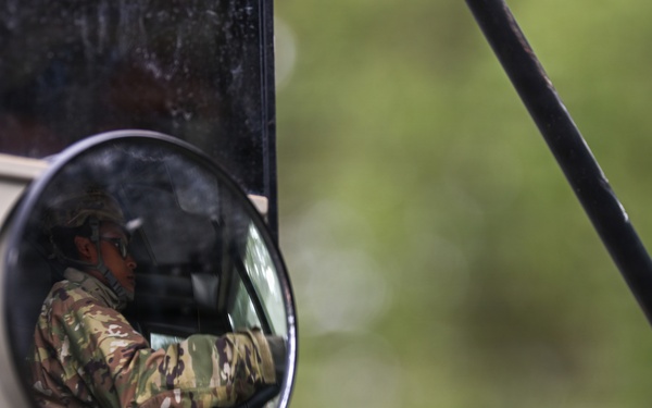 A person wearing military camouflage uniform and a cap is visible reflected in a vehicle's side mirror assembly. The reflection shows the individual seated inside a vehicle, appearing to be driving or operating it. The image is composed as a close-up of the mirror hardware against a blurred green background.