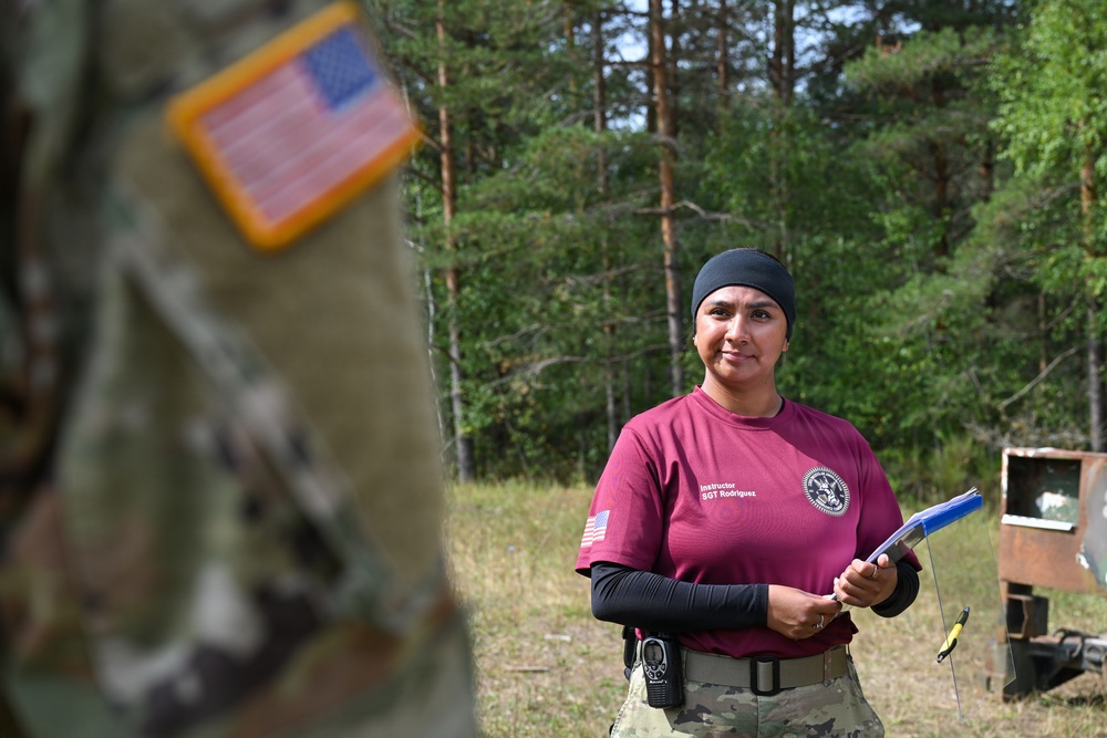 A U.S. Army instructor identified as SGT Rodriguez is standing outdoors in a wooded training area, wearing a maroon instructor t-shirt over a black long-sleeve undershirt and OCP (Operational Camouflage Pattern) pants. She is holding a clipboard and appears to be overseeing or evaluating a training exercise. In the foreground, a blurred soldier in full OCP uniform with a reverse U.S. flag patch is visible.