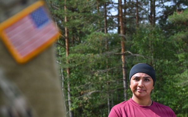 A U.S. Army instructor identified as SGT Rodriguez is standing outdoors in a wooded training area, wearing a maroon instructor t-shirt over a black long-sleeve undershirt and OCP (Operational Camouflage Pattern) pants. She is holding a clipboard and appears to be overseeing or evaluating a training exercise. In the foreground, a blurred soldier in full OCP uniform with a reverse U.S. flag patch is visible.