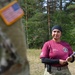 A U.S. Army instructor identified as SGT Rodriguez is standing outdoors in a wooded training area, wearing a maroon instructor t-shirt over a black long-sleeve undershirt and OCP (Operational Camouflage Pattern) pants. She is holding a clipboard and appears to be overseeing or evaluating a training exercise. In the foreground, a blurred soldier in full OCP uniform with a reverse U.S. flag patch is visible.