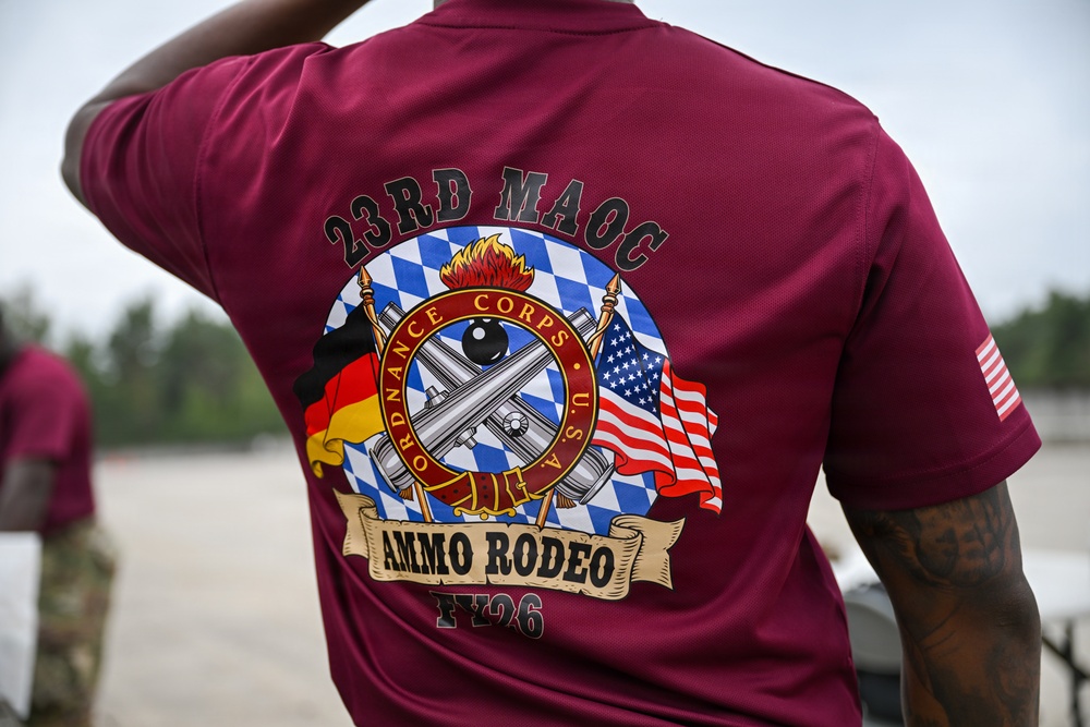The image shows the back of a maroon/burgundy athletic shirt worn by a person, featuring a detailed military unit graphic. The shirt belongs to the 23rd MAOC (Munitions and Armaments Operations Company) and commemorates an 'Ammo Rodeo' event for FY26. The design incorporates the U.S. Army Ordnance Corps insignia flanked by German and American flags against a Bavarian blue-and-white diamond pattern background.