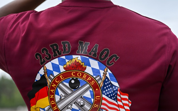 The image shows the back of a maroon/burgundy athletic shirt worn by a person, featuring a detailed military unit graphic. The shirt belongs to the 23rd MAOC (Munitions and Armaments Operations Company) and commemorates an 'Ammo Rodeo' event for FY26. The design incorporates the U.S. Army Ordnance Corps insignia flanked by German and American flags against a Bavarian blue-and-white diamond pattern background.